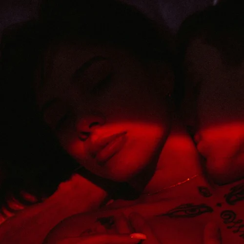 Casal em momento íntimo sob iluminação vermelha intensa, mulher com tatuagens deitada sobre o parceiro, sem rostos visíveis